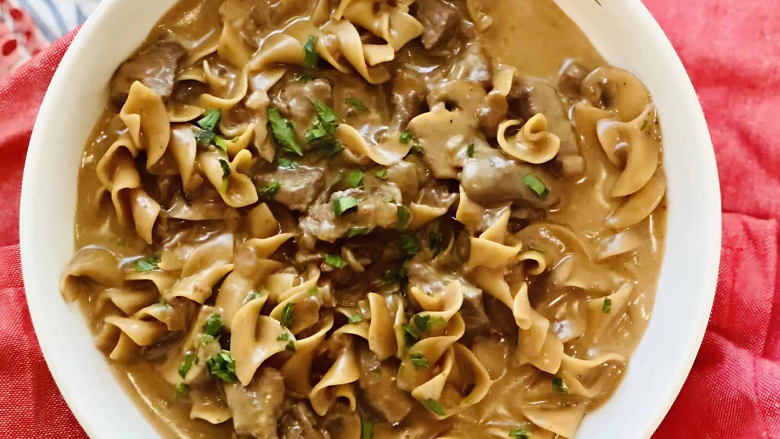 A bowl of beef stroganoff with noodles, topped with fresh parsley. The dish features a creamy sauce mixed with pieces of beef and wide, curly noodles, set on a red cloth background.