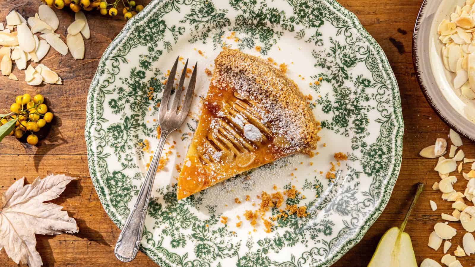 A slice of fruit tart with powdered sugar sits on a decorative plate beside a fork and scattered almond slices on a wooden table.