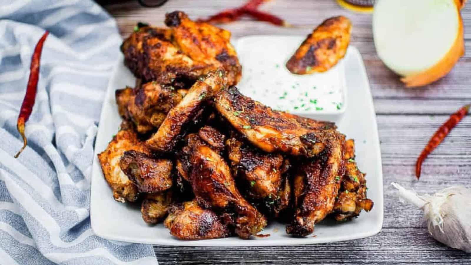 A plate of grilled chicken wings with a side of creamy dipping sauce. The dish is garnished with herbs and surrounded by chili peppers, an onion slice, and garlic on a wooden surface. A striped cloth is draped nearby.