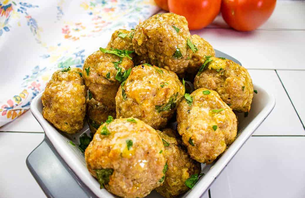 A bowl of golden-brown meatballs garnished with fresh herbs. The dish is placed on a white and gray square plate. In the background, there's a floral-patterned cloth and a pair of ripe tomatoes. The setting is on a white tiled surface.