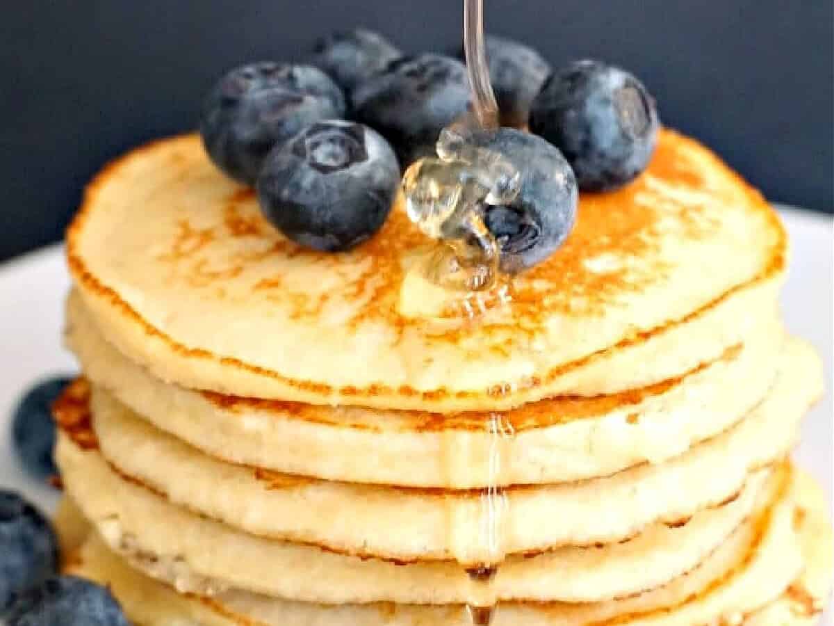 A stack of golden pancakes topped with fresh blueberries and drizzled with honey, set on a white plate against a dark background.