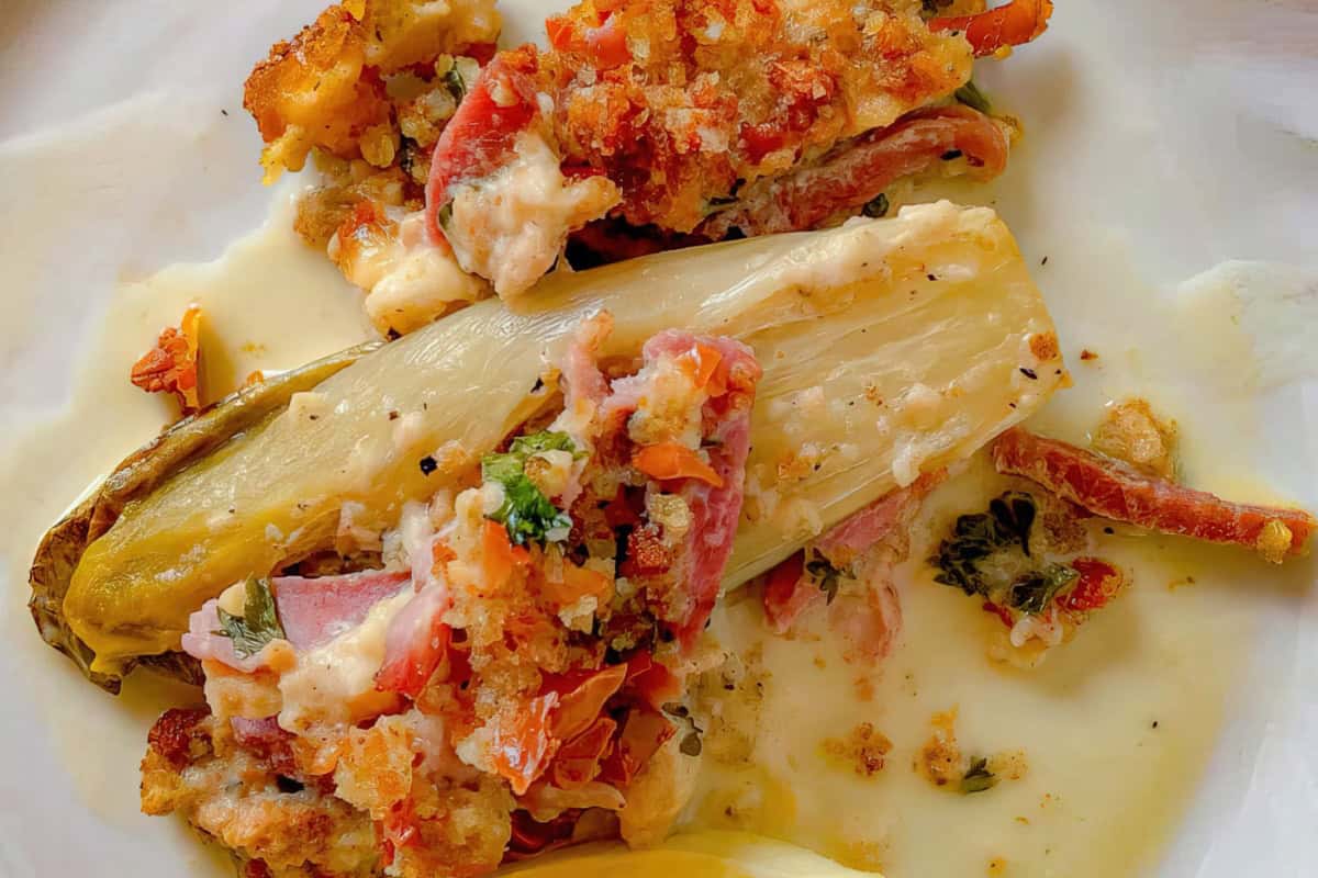 A plate featuring endive stuffed with a mixture of breadcrumbs, tomatoes, herbs, and melted cheese. The dish is covered in a creamy sauce, creating a rich and appetizing presentation.