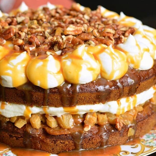 A layered cake topped with whipped cream, caramel sauce, and chopped pecans; visible filling of cream and caramelized apples.