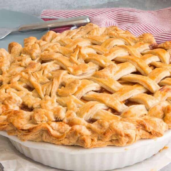 A baked pie with a golden brown lattice crust in a white dish sits beside stacked plates, utensils, and a red-striped cloth.