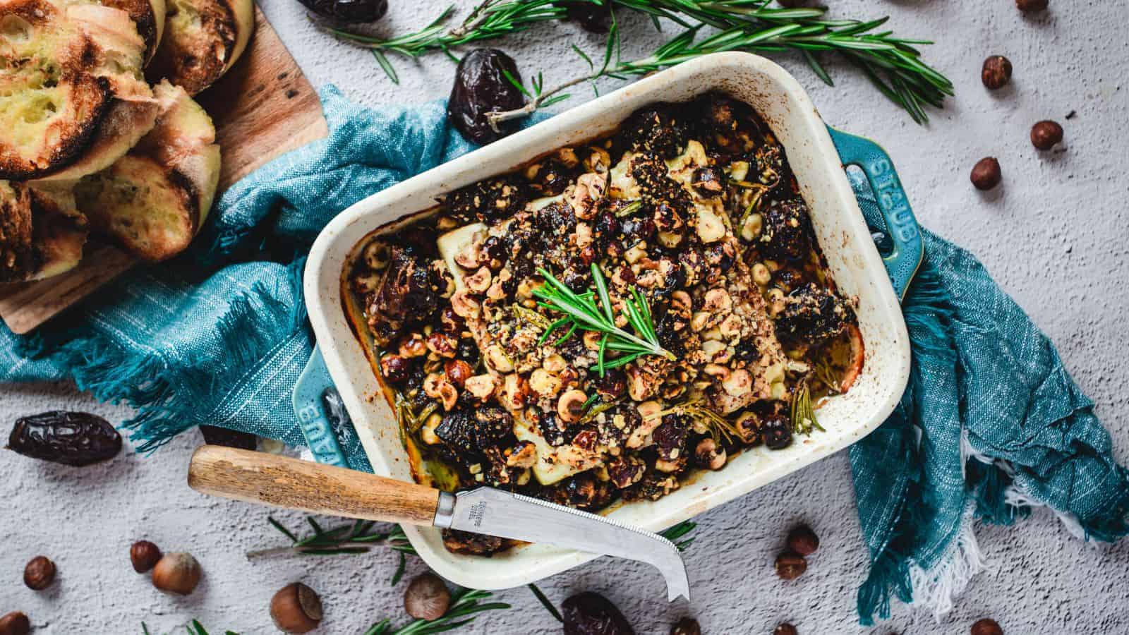 A casserole dish filled with a baked dish topped with herbs and nuts, surrounded by rosemary sprigs, dates, and hazelnuts. A knife rests on the dish's edge. To the side, a wooden board holds slices of toasted bread.