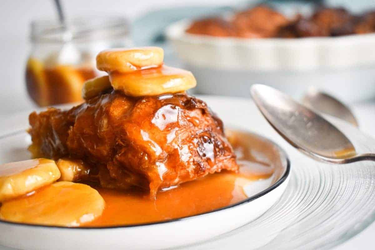A dessert featuring a slice of glazed cake topped with banana slices and caramel sauce on a white plate. A spoon is placed beside the plate, and a jar of caramel is in the background.