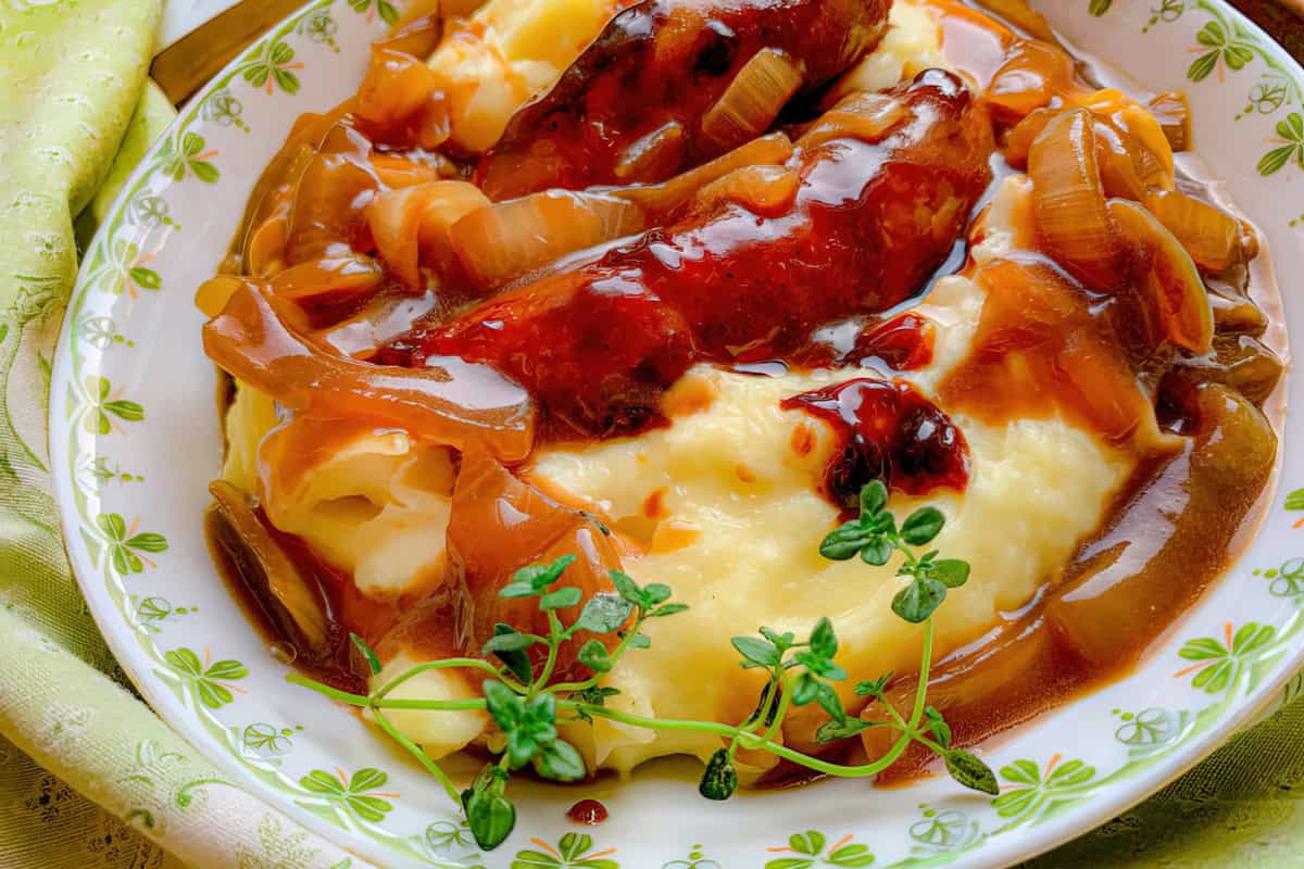 The image shows a plate featuring sausages with caramelized onions in gravy, served over mashed potatoes. The dish is garnished with fresh thyme.