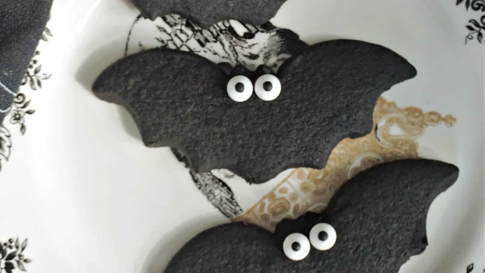 Three black bat-shaped cookies with white candy eyes are on a decorative white plate. The plate features intricate black and gold designs.