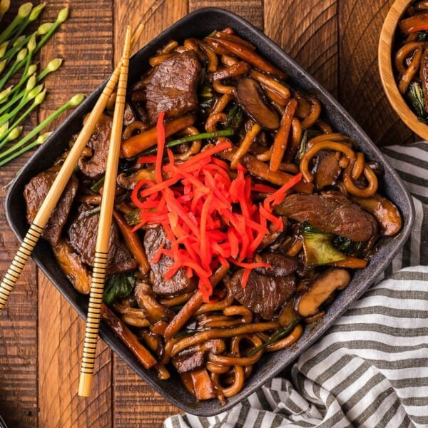 A bowl of stir-fried noodles with beef, mushrooms, vegetables, and shredded red pickled ginger, with chopsticks on a striped cloth.
