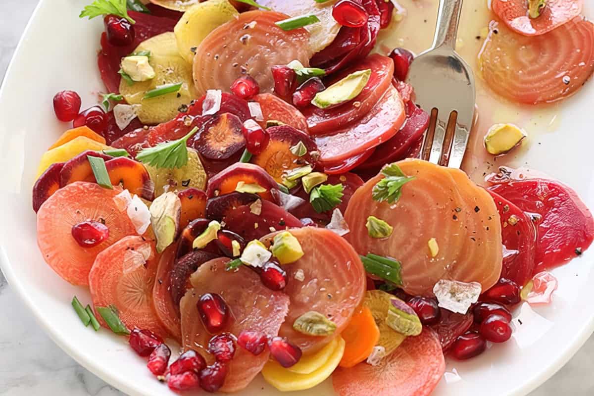 A colorful beet salad on a white plate, featuring sliced red and yellow beets, pomegranate seeds, pistachios, and fresh herbs. A fork rests on the side, and the salad is lightly dressed and sprinkled with cracked pepper.