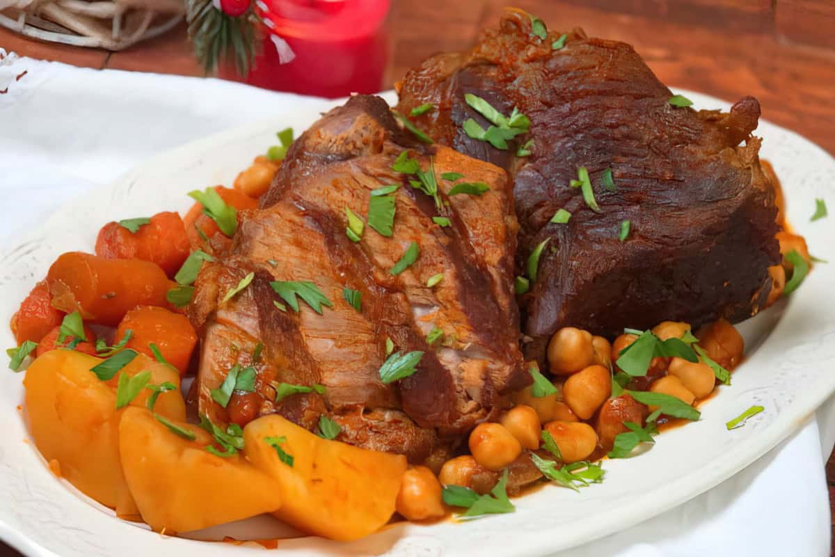 A plate of succulent roasted meat garnished with fresh parsley, surrounded by cooked chickpeas, carrots, and potatoes. A red candle and festive decor are visible in the blurred background.