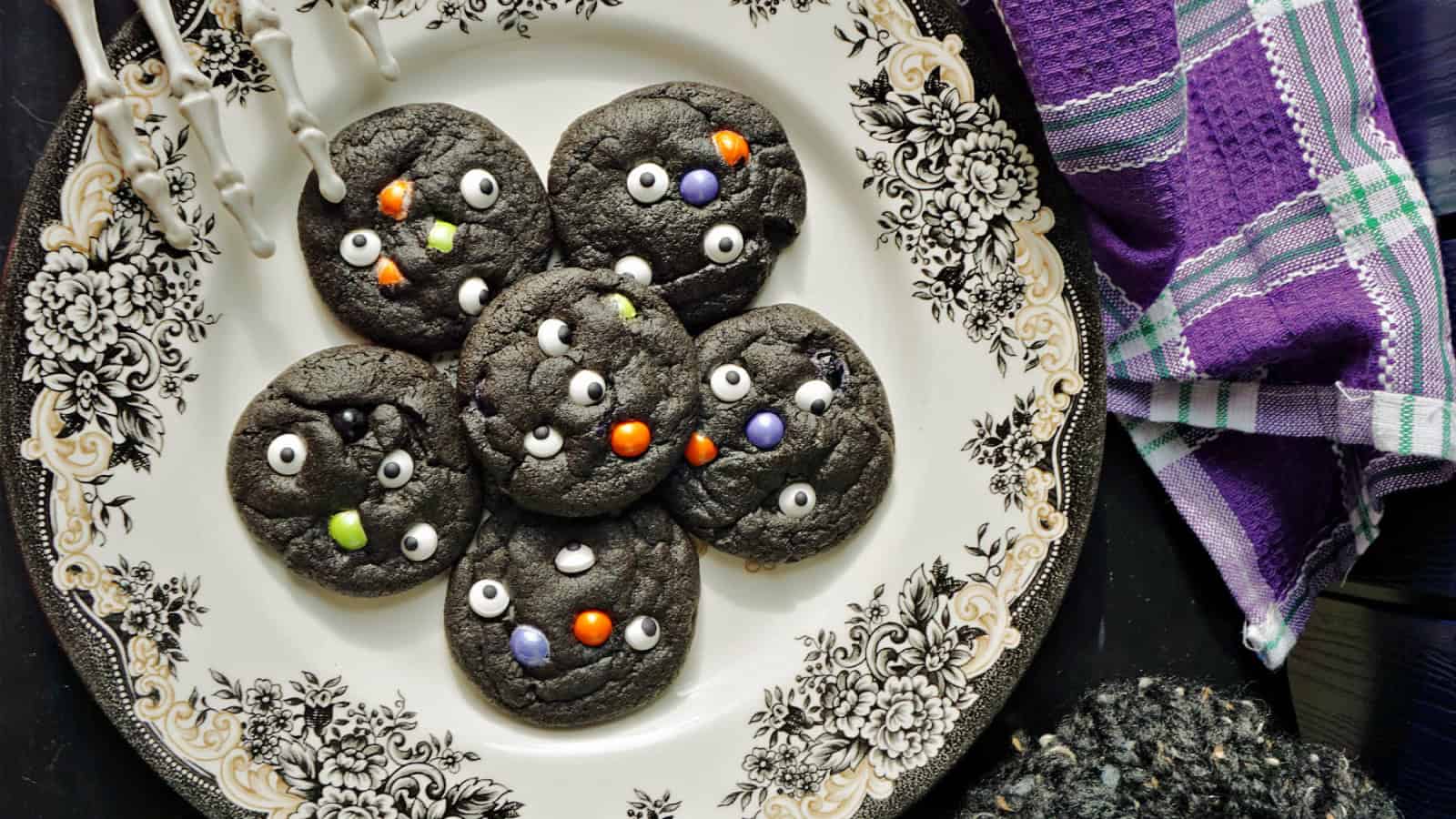 A vintage plate holds six black cookies decorated with small white candy eyes and colorful candies. A purple plaid kitchen towel is nearby, along with a decorative fork with skeleton hands. The setting is spooky and festive.