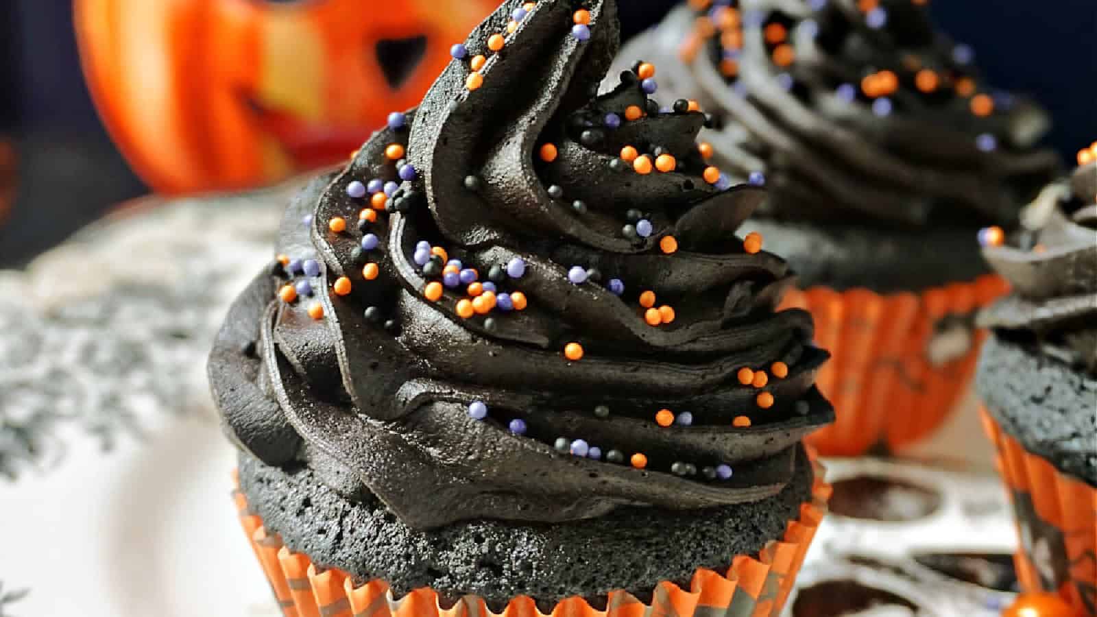 A close-up of Halloween-themed cupcakes with swirled black frosting topped with orange and purple sprinkles. The cupcakes are in orange wrappers, and a carved pumpkin decoration is blurred in the background.