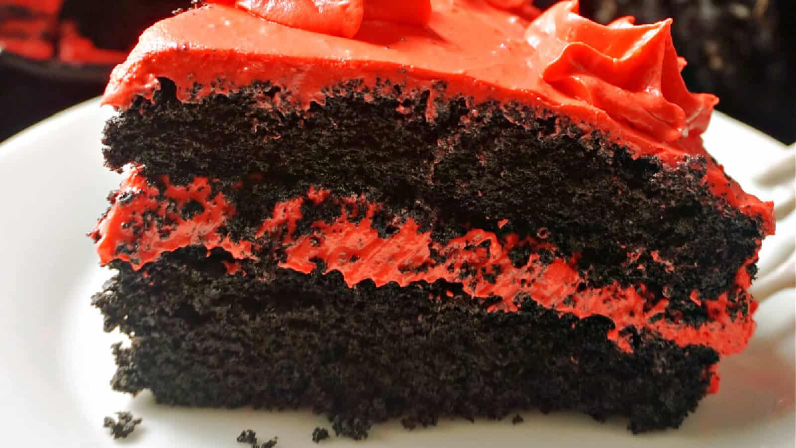 A close-up of a slice of rich, dark chocolate cake with vibrant red frosting. The cake appears moist and layered, with red frosting between each layer and decorative swirls on top, placed on a white plate. The background is blurred.