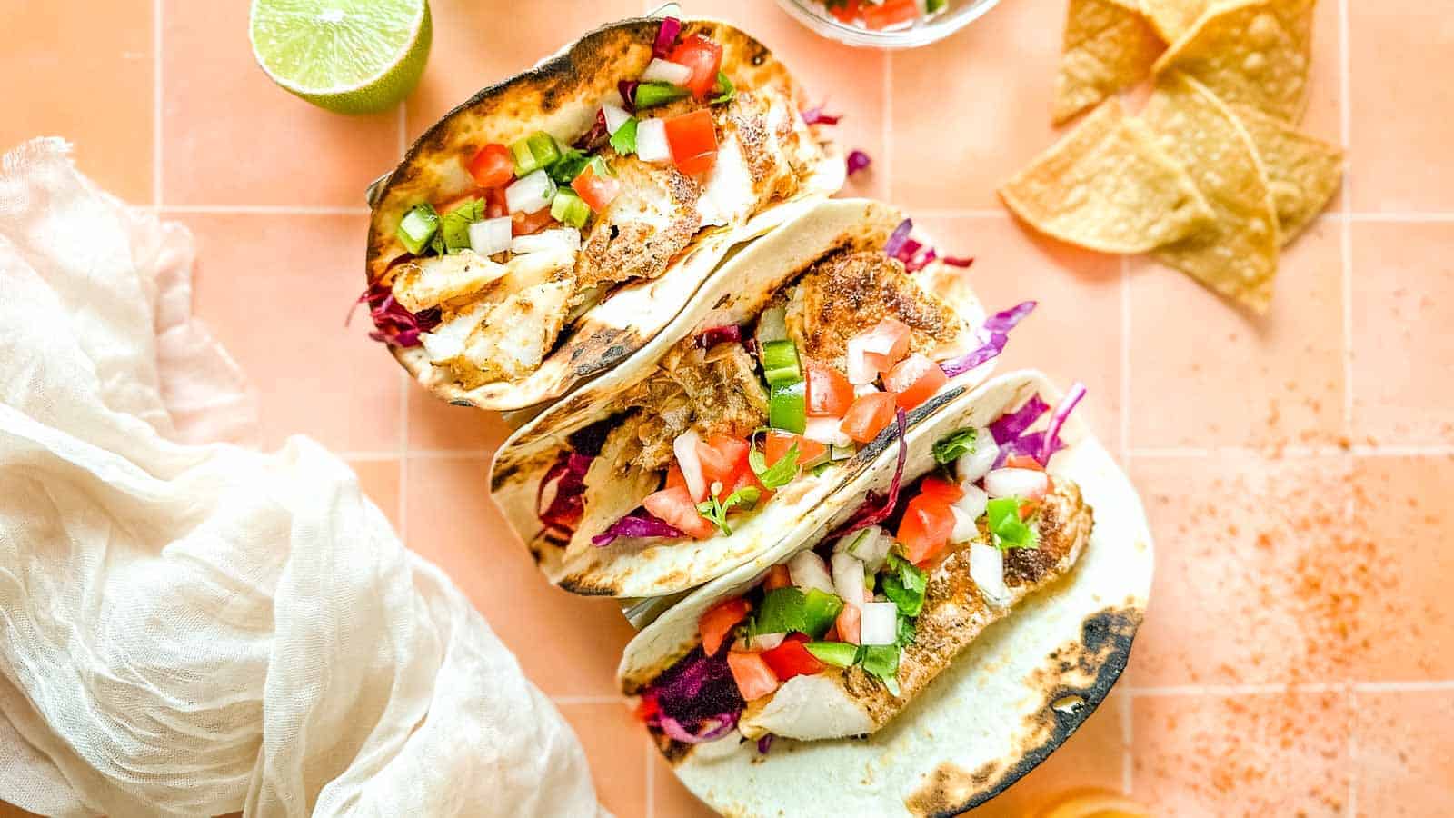 Three grilled fish tacos with fresh toppings like diced tomatoes, cucumbers, and purple cabbage are displayed on a tiled surface. Lime and tortilla chips are on the side, with a light cloth napkin nearby.