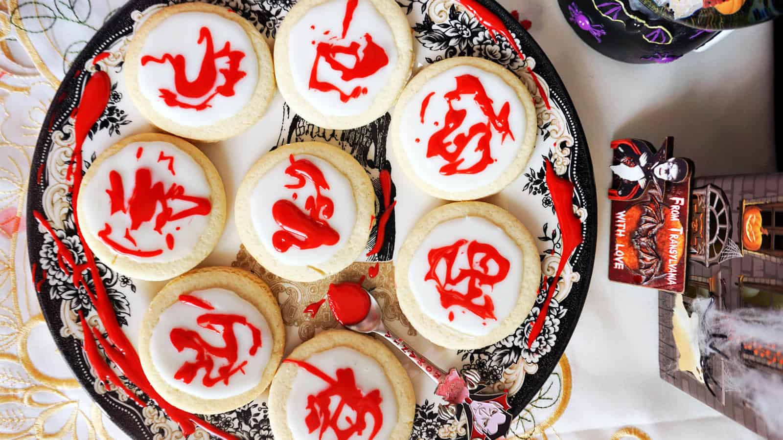 A decorative plate holds cookies with white icing, each featuring a red icing dragon symbol. The plate is set on a festive Halloween-themed tablecloth, accompanied by a small decorative coffin that reads, "From Transylvania With Love.