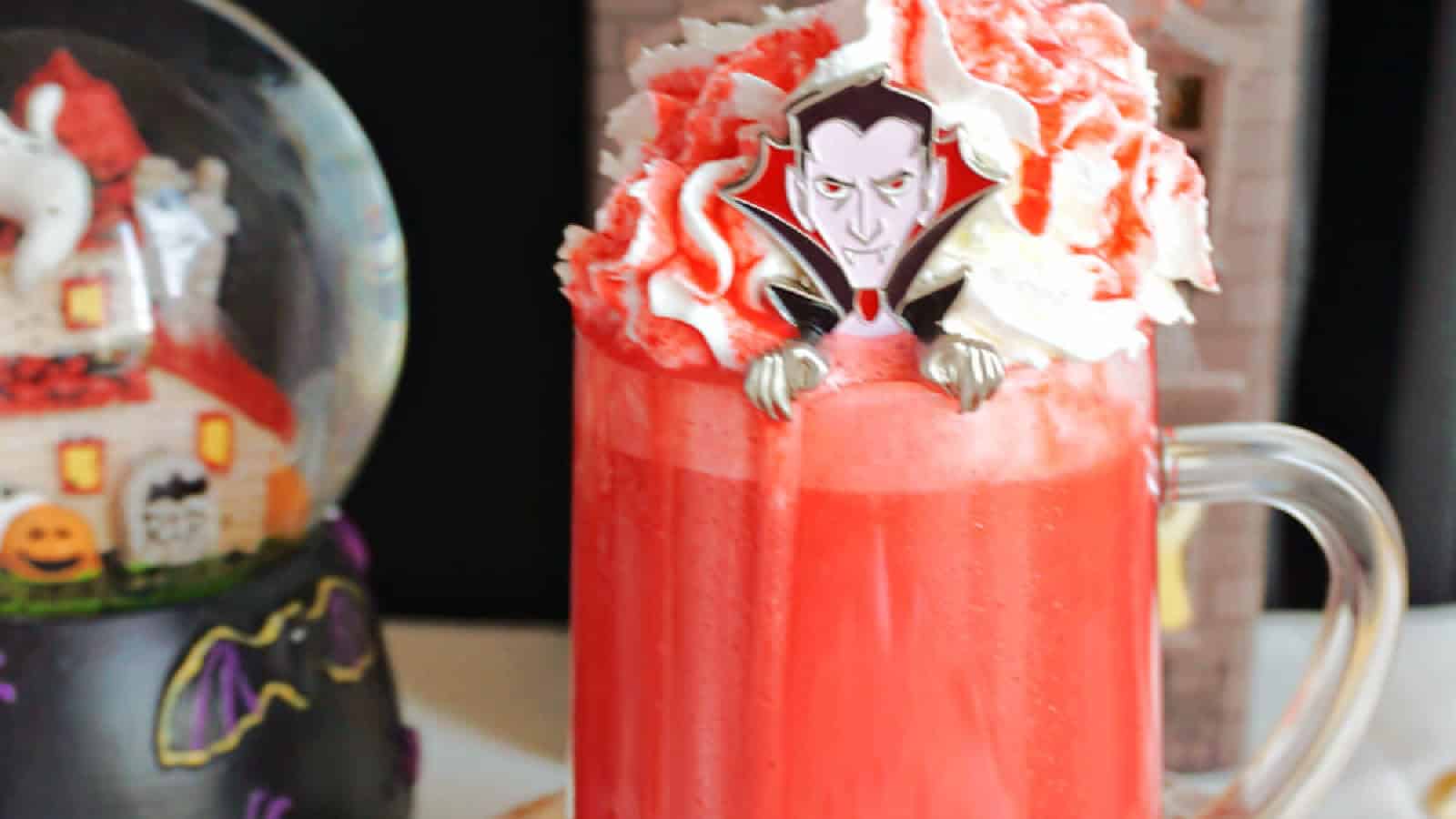 A glass mug filled with a red hot chocolate is topped with whipped cream and a Halloween-themed vampire decoration. In the background, there is a snow globe adorned with Halloween motifs.