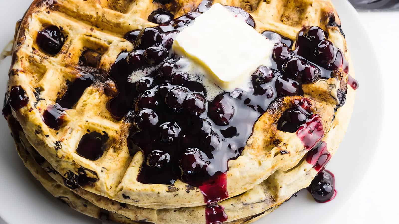 A stack of waffles topped with blueberry compote and a square of butter, served on a white plate. The vibrant blueberries and syrup cascade over the golden-brown waffles, creating a delicious and inviting breakfast scene.