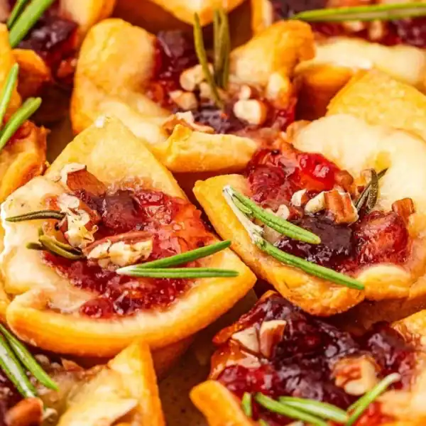 Mini pastry cups holding melted cheese, red jam, chopped nuts, and sprigs of fresh rosemary.