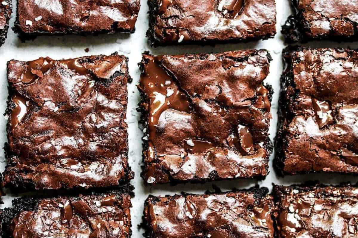Close-up of nine delicious, fudgy chocolate brownies arranged in a grid pattern on a parchment paper. The brownies have a shiny, cracked top and bits of melted chocolate visible, making them look rich and indulgent.
