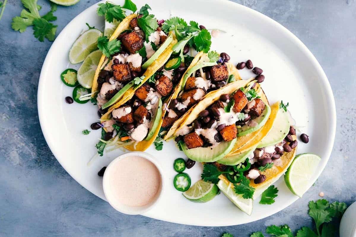 A plate of tacos filled with crispy tofu, black beans, avocado slices, and fresh cilantro, accompanied by lime wedges and sliced jalapeños. A small bowl of dipping sauce is placed beside the tacos on a gray background.