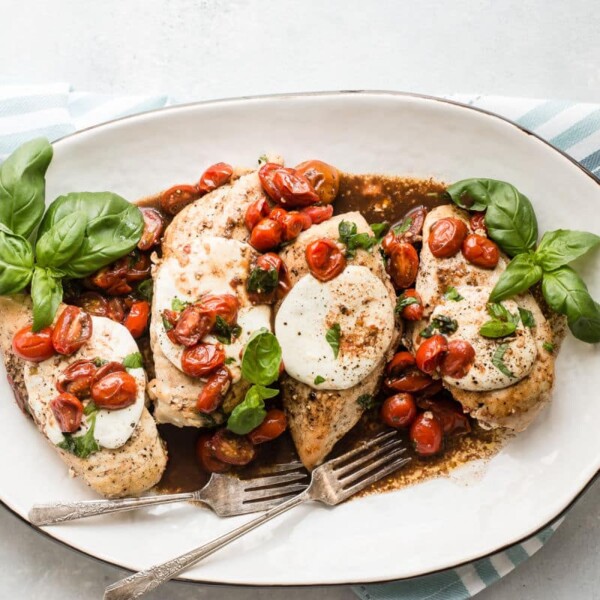 Chicken breast pieces with mozzarella, cherry tomatoes, and basil on a white platter with two forks on a striped cloth.