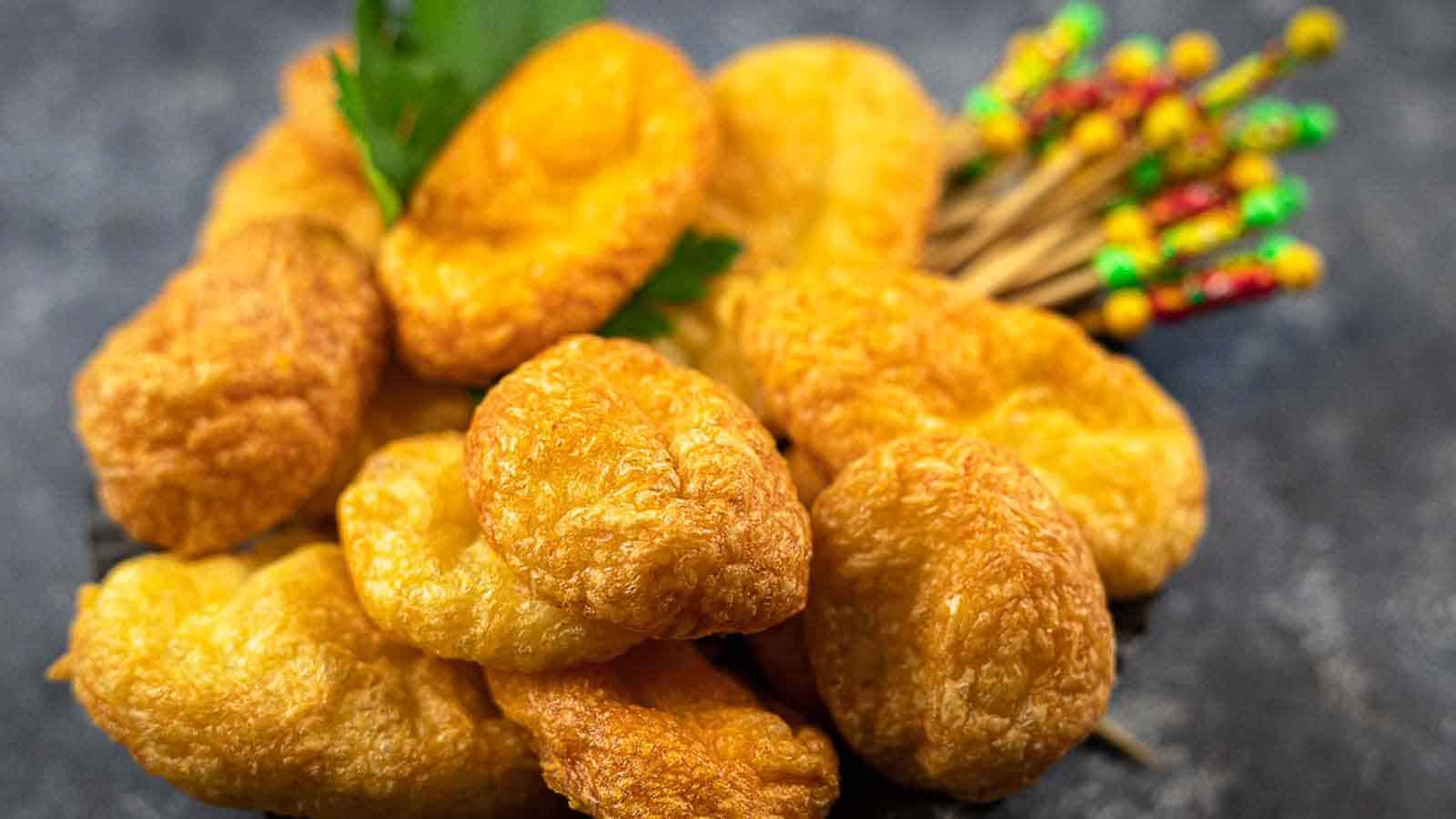 A pile of golden brown crunchy snacks, resembling fried dumplings or pastries, is stacked on a dark surface. A small bunch of colorful toothpicks and a garnish of parsley accompany the snacks.