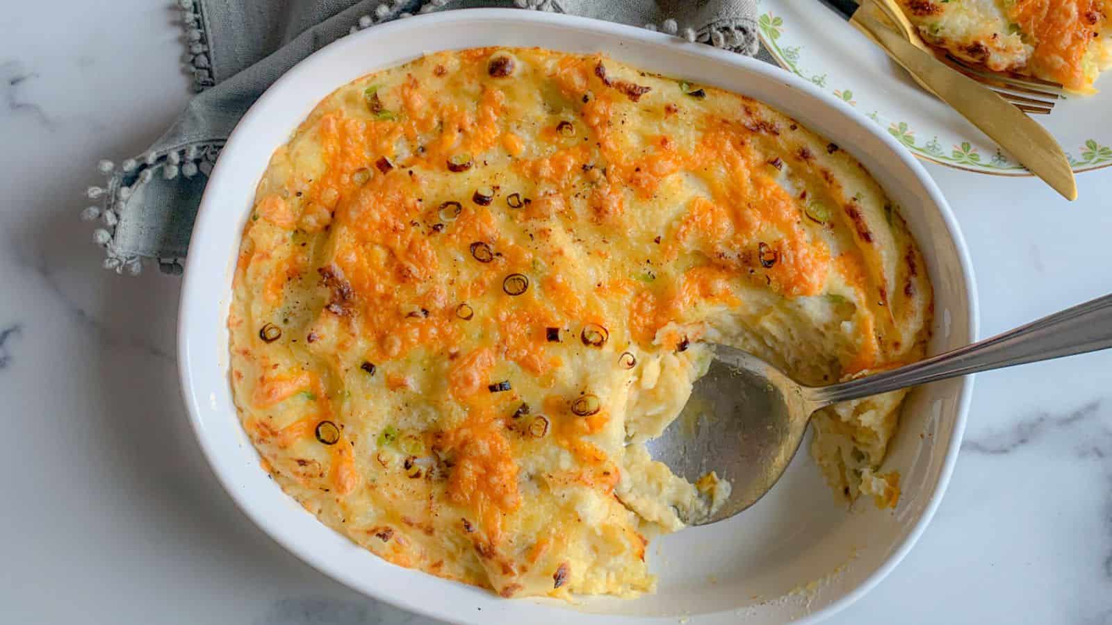 The image shows baked mashed potatoes in a white oval dish with a serving spoon. The top is covered with melted cheese and garnished with chopped green onions. Nearby, a piece of the dish is served on a plate.