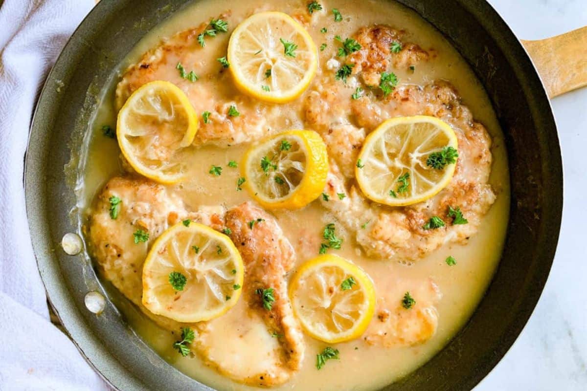 Chicken breasts in lemon sauce with lemon slices and chopped parsley, served in a skillet.