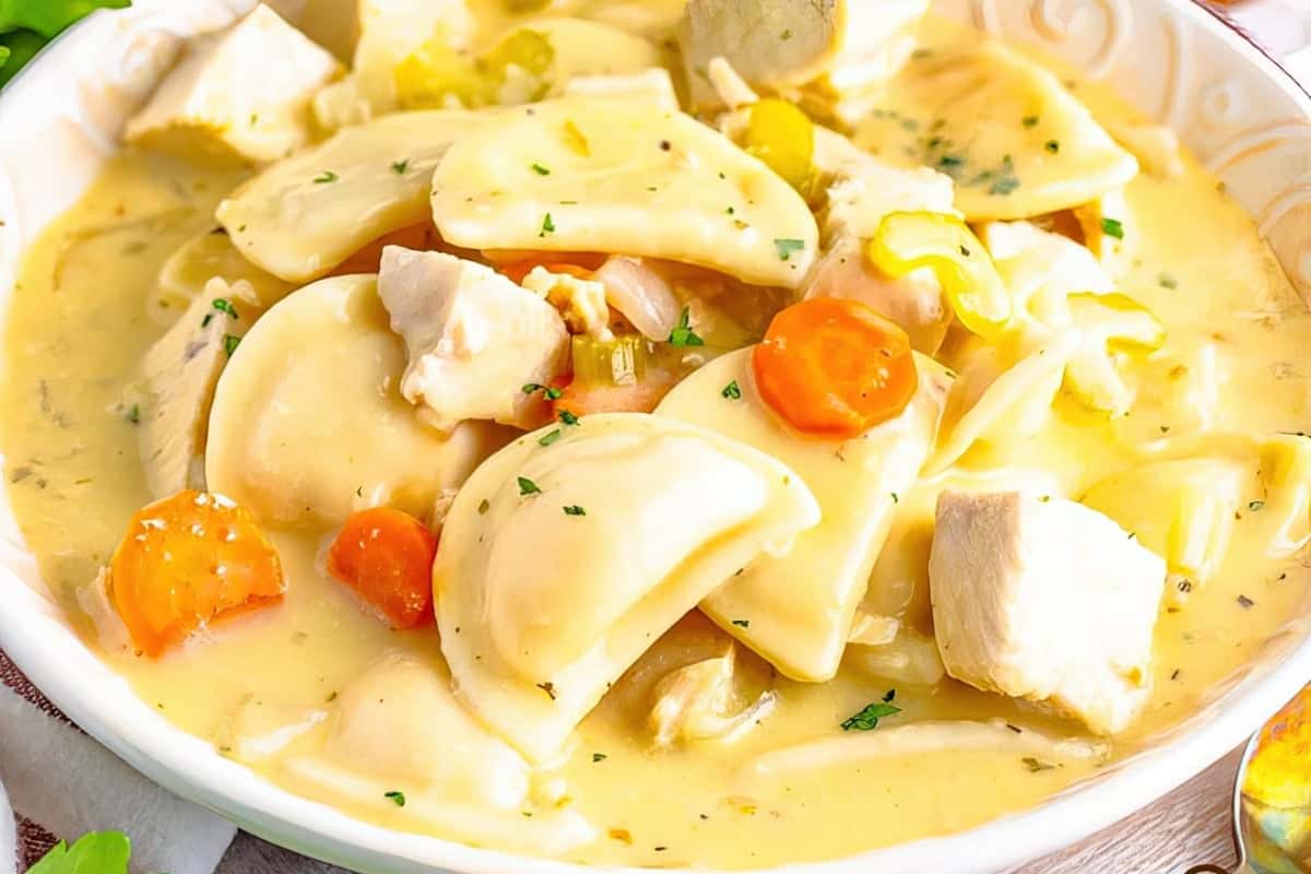 A creamy soup filled with thick noodles, chunks of chicken, sliced carrots, and celery in a white bowl. The dish is garnished with chopped herbs and has a rich, hearty appearance.