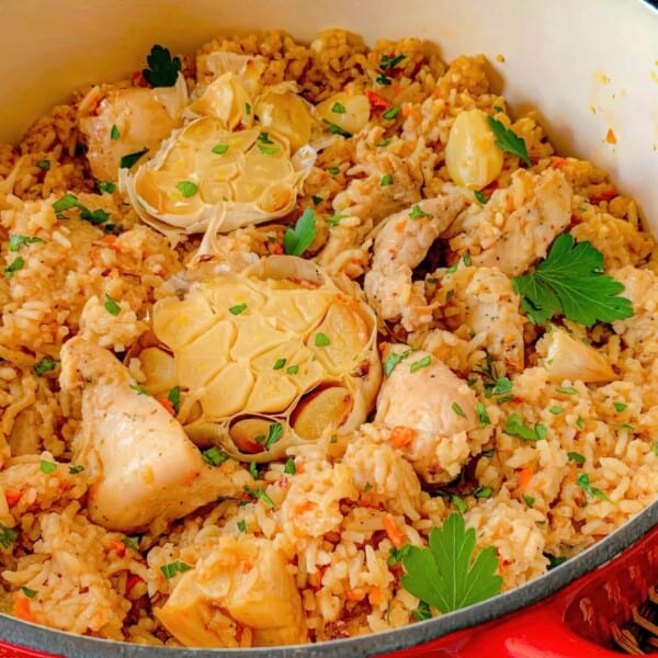 A pot filled with cooked rice, chicken pieces, whole roasted garlic bulbs, and parsley garnish.