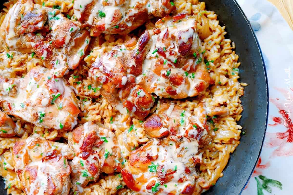 A skillet filled with cooked rice topped with seasoned chicken pieces. The dish is garnished with fresh herbs and drizzled with a creamy sauce.