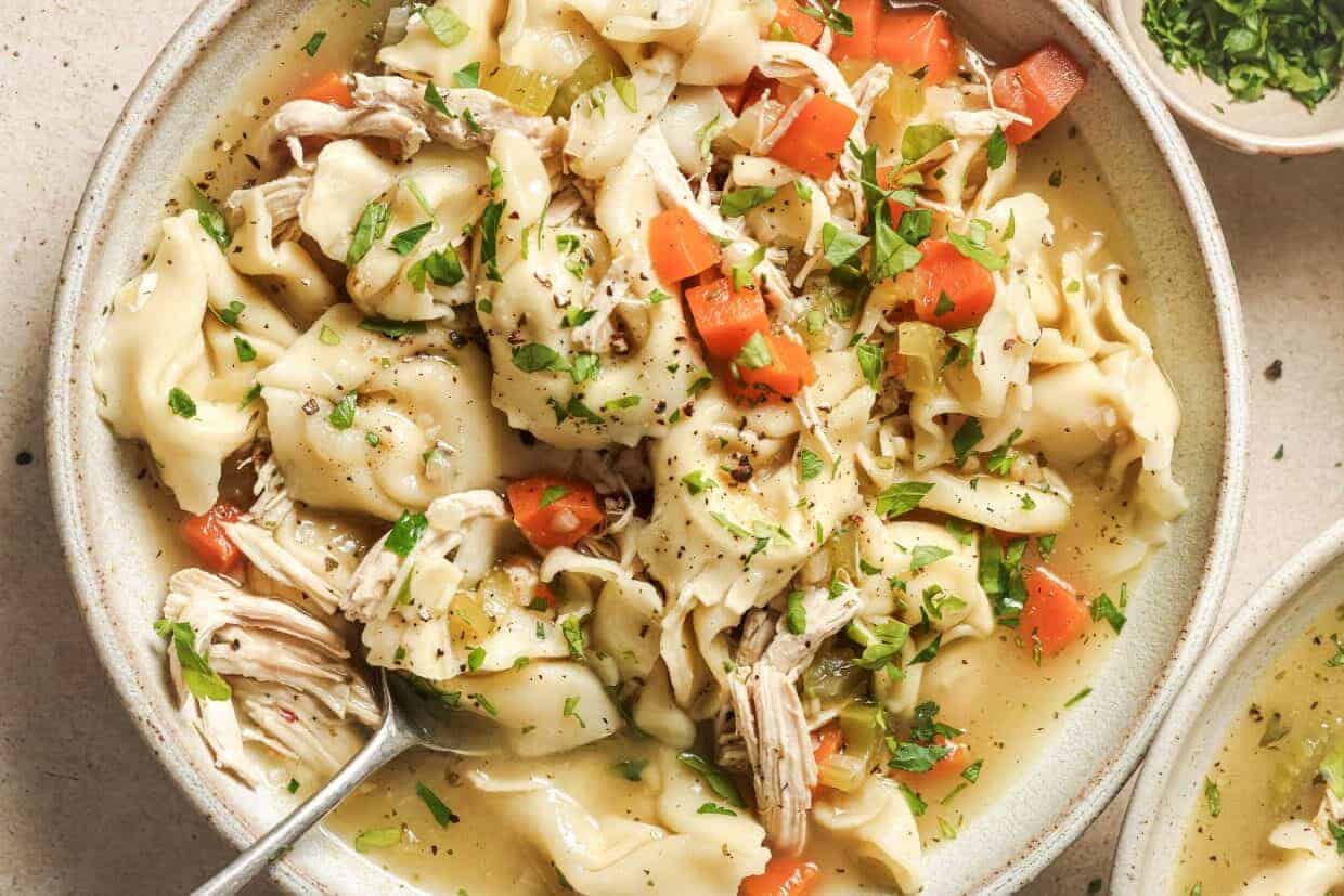 A bowl of tortellini soup filled with shredded chicken, diced carrots, and celery in a savory broth. Garnished with fresh parsley and cracked black pepper, it sits on a light-colored surface. A spoon rests in the bowl, ready for serving.