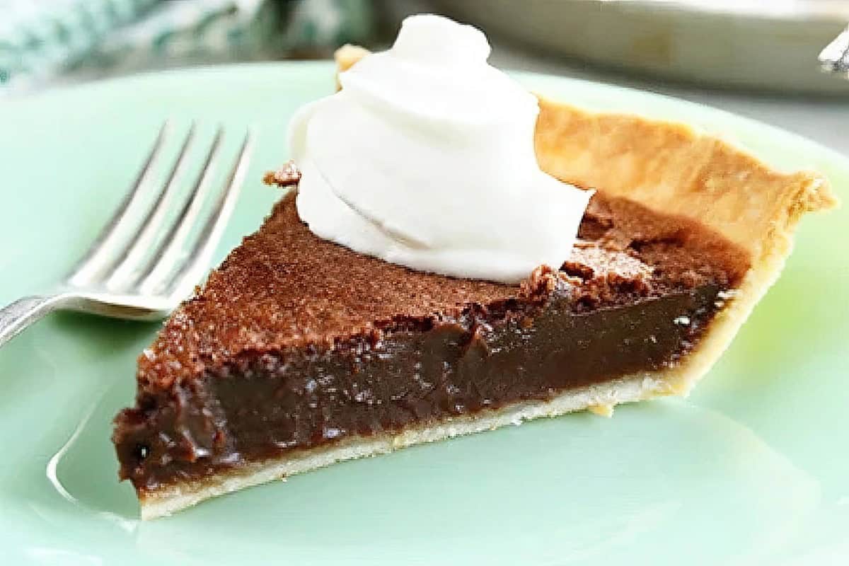 A slice of chocolate pie topped with a dollop of whipped cream sits on a light green plate. A fork is placed next to the slice. The flaky crust and rich, glossy chocolate filling are visible in the pie.