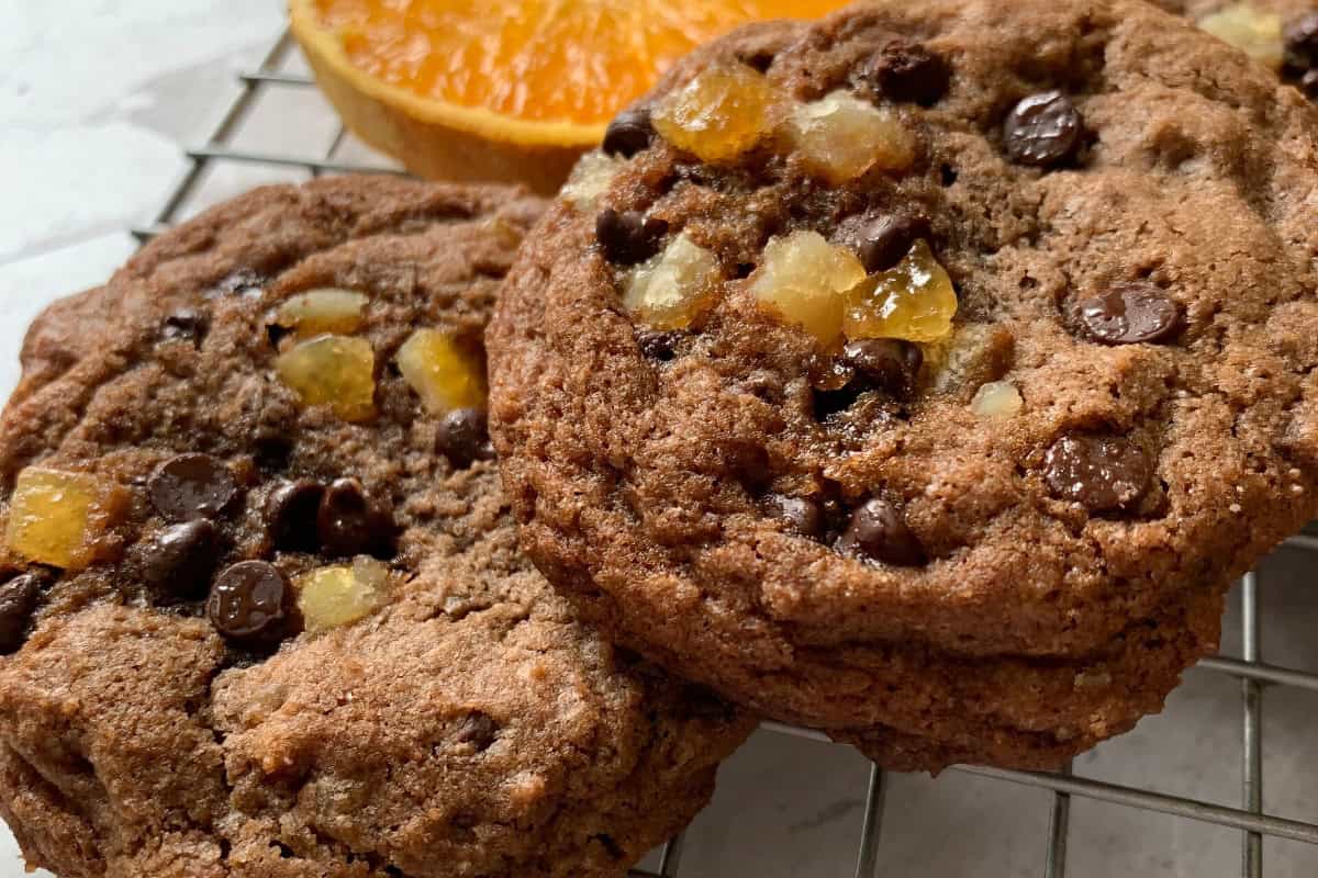Chocolate chip cookies with chunks of candied orange rest on a cooling rack. An orange slice is visible in the background.