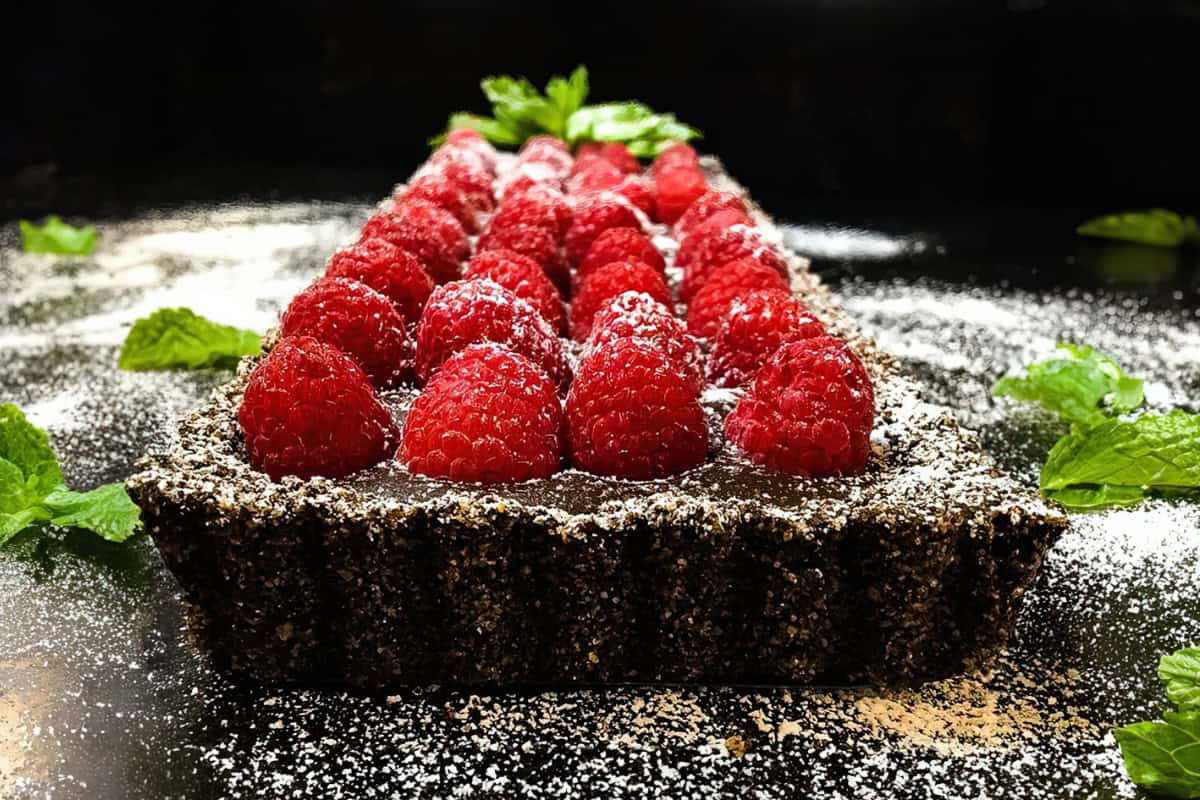 A rectangular chocolate tart topped with fresh raspberries is displayed on a dark surface. Mint leaves surround the tart, and powdered sugar is sprinkled around, adding a touch of elegance.