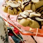 Christmas almond cookies with chocolate drizzle and sliced almonds on a plate beside a decorative teapot and colorful cloth.