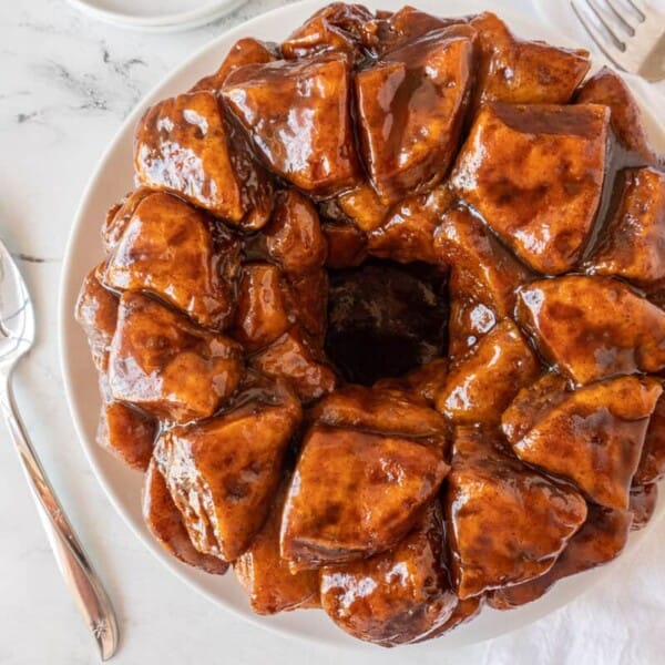 A glazed monkey bread ring on a white plate, with a serving spoon and forks nearby on a marble surface.