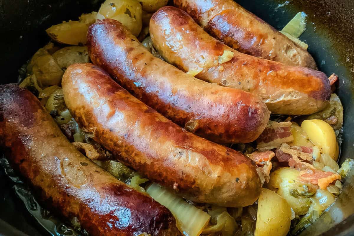 Five browned sausages on top of sliced potatoes, onions, and pieces of bacon in a pan.