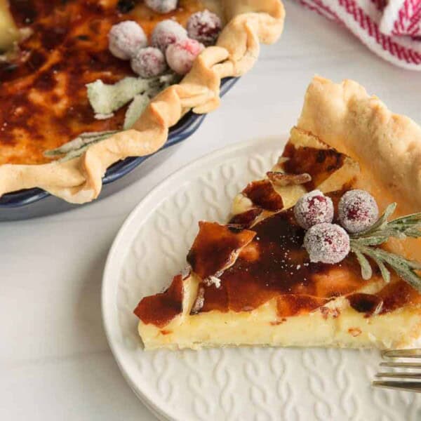 A slice of custard pie garnished with sugared cranberries and herbs sits on a white plate, with the rest of the pie and a fork nearby.