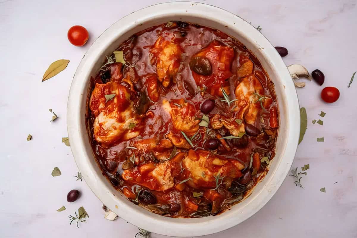 A round dish filled with baked chicken thighs in a rich tomato sauce, garnished with olives, herbs, and mushrooms. Scattered around are cherry tomatoes, bay leaves, and olives on a light background.