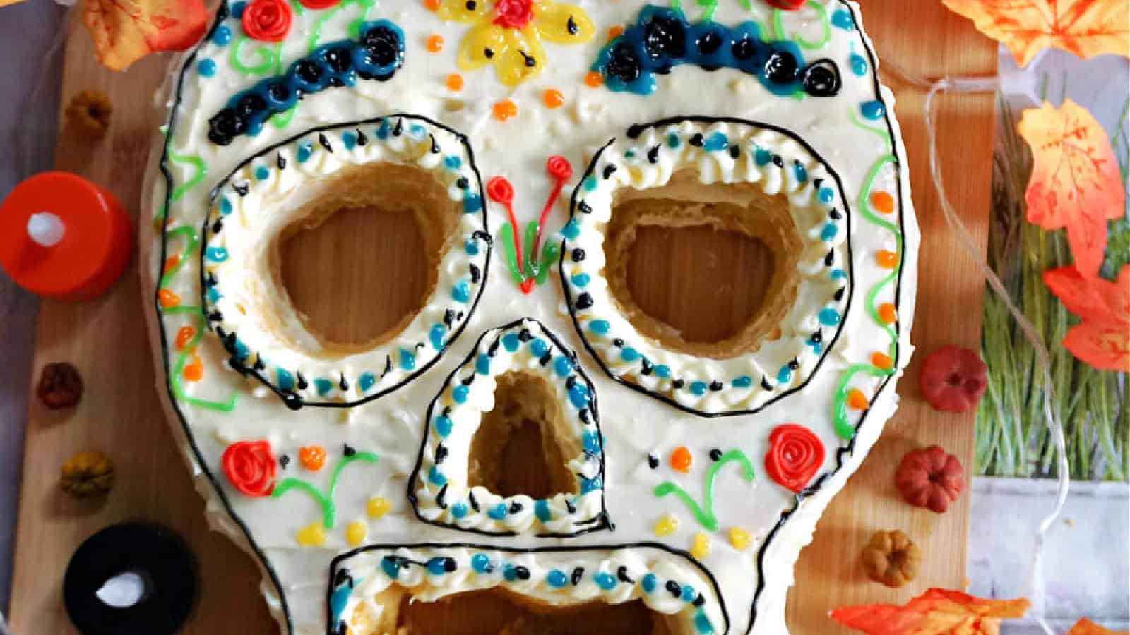 A colorful sugar skull-shaped cake decorated with icing patterns in various colors, including blue, orange, and yellow. It is surrounded by small pumpkins and autumn leaves on a wooden surface.