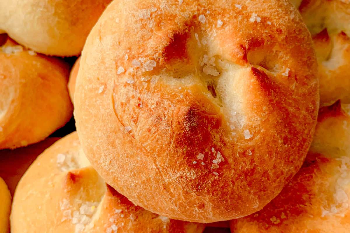 Close-up of freshly baked, golden brown bread rolls with a lightly crisp crust and a sprinkling of coarse salt on top. The rolls are piled together showcasing their warm, appetizing texture.