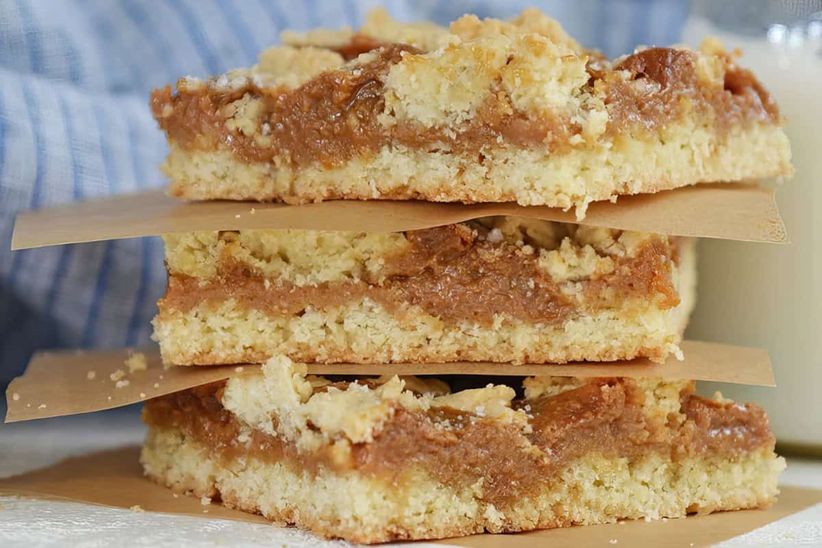 Three stacked caramel crumb bars with a crumbly topping and creamy filling are separated by parchment paper. They rest on a surface with a checkered blue background visible.