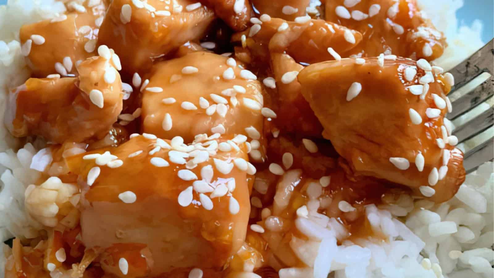 A close-up of a delicious dish featuring bite-sized pieces of chicken glazed in a sticky orange sauce, garnished with white sesame seeds, served over fluffy white rice. A fork is placed to the side.