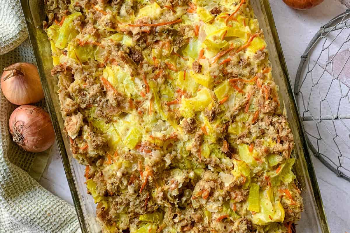 A baking dish with a golden-brown casserole topped with bread pieces and vegetables. Whole onions and a kitchen towel sit beside it.