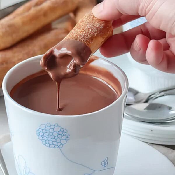 A hand dips a churro into a cup of thick hot chocolate, with more churros and utensils on a plate in the background.