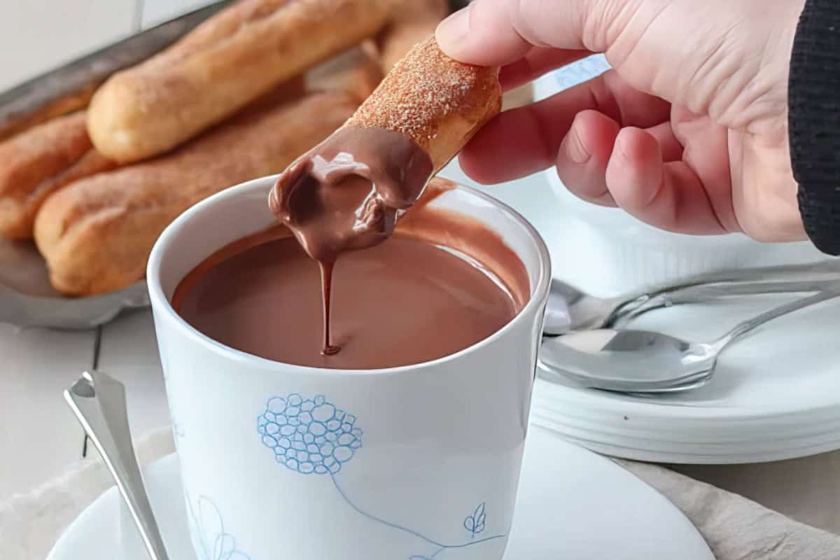 A hand dips a churro covered in sugar into a cup of thick hot chocolate. In the background, a tray holds more churros. Two spoons rest beside the cup on a white surface. The cup has a blue floral design.