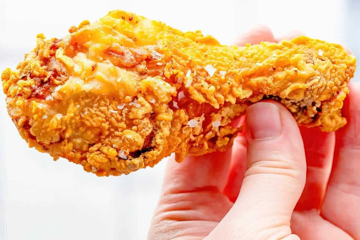 Close-up of a hand holding a crispy, golden fried chicken drumstick against a light background.