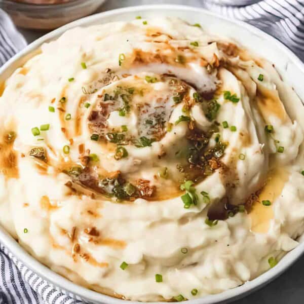 A bowl of mashed potatoes with melted butter, brown gravy, and chopped chives sits on a table next to a striped cloth.